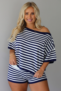 A woman wearing a navy and white horizontally striped off-the-shoulder top and matching striped shorts, standing indoors against a white wall.