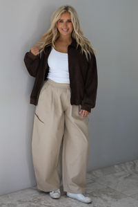 A woman wearing a brown suede jacket with a white tank and sand colored baggy pants, standing indoors against a white wall with white marble flooring. 