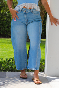 A woman wearing light-wash blue cropped barrel cut jeans with an asymmetrical button closure, standing outdoors on a patio with green grass and plants in the background.