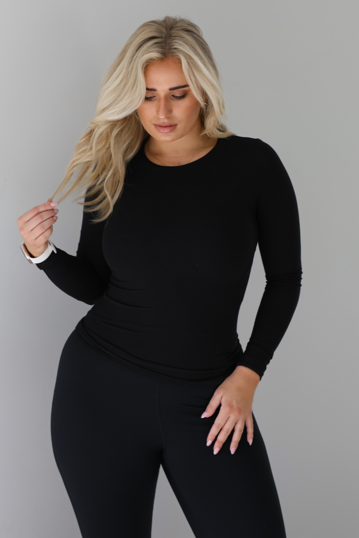 Woman wearing a black, high neckline, long sleeved fitted top that has a ribbed design to the material. It's paired with black leggings. The photo is taken against a white wall. 