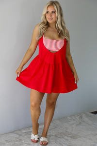 Woman wearing a red dress with a red and white gingham romper underneath. The photo is taken indoors against a white wall with white marble flooring. 