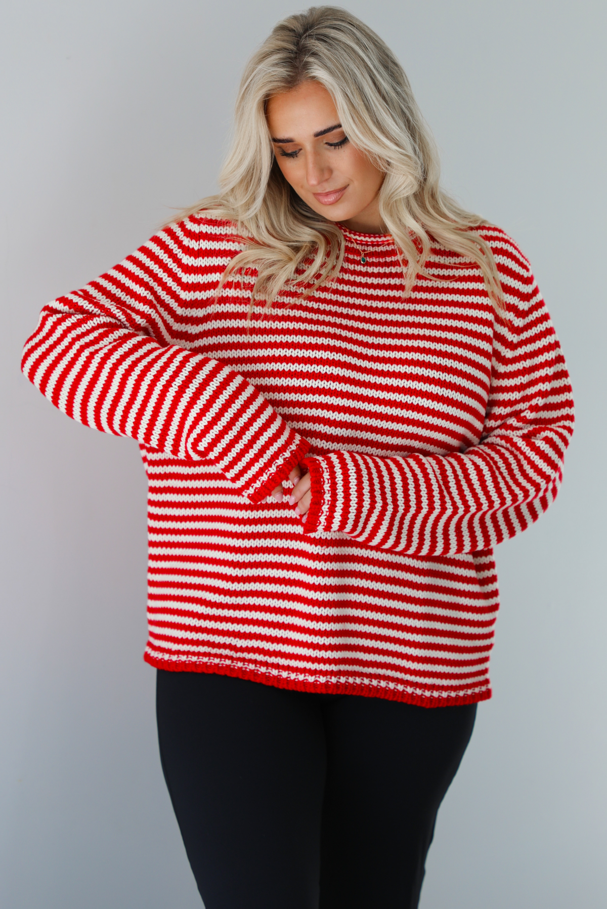 Woman wearing a white and red striped sweater that has a high neckline, an oversized fit, and is paired with black leggings. The photo shows a closeup of the sweater. The photo is taken indoors against a white wall.