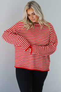 Woman wearing a white and red striped sweater that has a high neckline, an oversized fit, and is paired with black leggings. The photo shows a closeup of the sweater. The photo is taken indoors against a white wall.