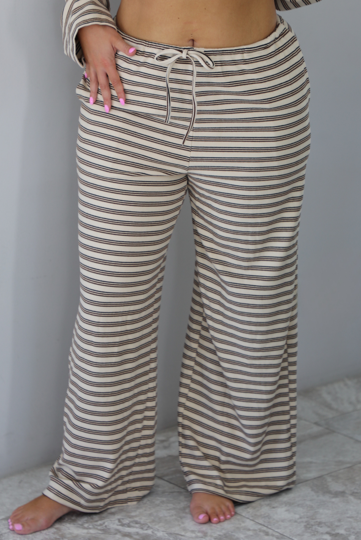 A woman shown from the waist down wearing a sand colored matching two piece set with a brown striped pattern throughout featuring an oversized long sleeved top and oversized slouchy wide leg pants, standing indoors against a white wall with white marble flooring. The photo focuses on the pants, which have an elastic and drawstring waistline and functional pockets on either side. 