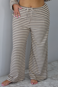 A woman shown from the waist down wearing a sand colored matching two piece set with a brown striped pattern throughout featuring an oversized long sleeved top and oversized slouchy wide leg pants, standing indoors against a white wall with white marble flooring. The photo focuses on the pants, which have an elastic and drawstring waistline and functional pockets on either side. 