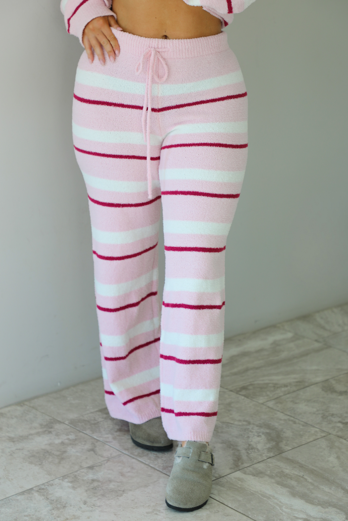 Woman wearing a pink set with a darker pink and white striped design. The set consists of an oversized sweater top and high waisted pants. The photo shows a closeup of the bottoms. The photo is taken indoors against a white wall with white marble flooring. 