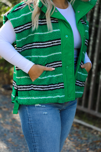 Woman model wearing a green, black, and white striped vest that has a sherpa texture to it, functional button closures in the front, and tie closures on either side, It is paired with a white long sleeved top, a medium wash pair of denim jeans, and tan ugg boots. The photo is taken outdoors with a metal gate and greenery in the back. The vest is shown close up on the side, displaying the unclasped buttons, the pockets in use, and the tie details on either side.