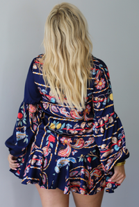 A woman shown from behind modeling a navy blue mini dress with a colorful floral print and long balloon sleeves, paired with beige ankle boots, standing indoors against a white wall.