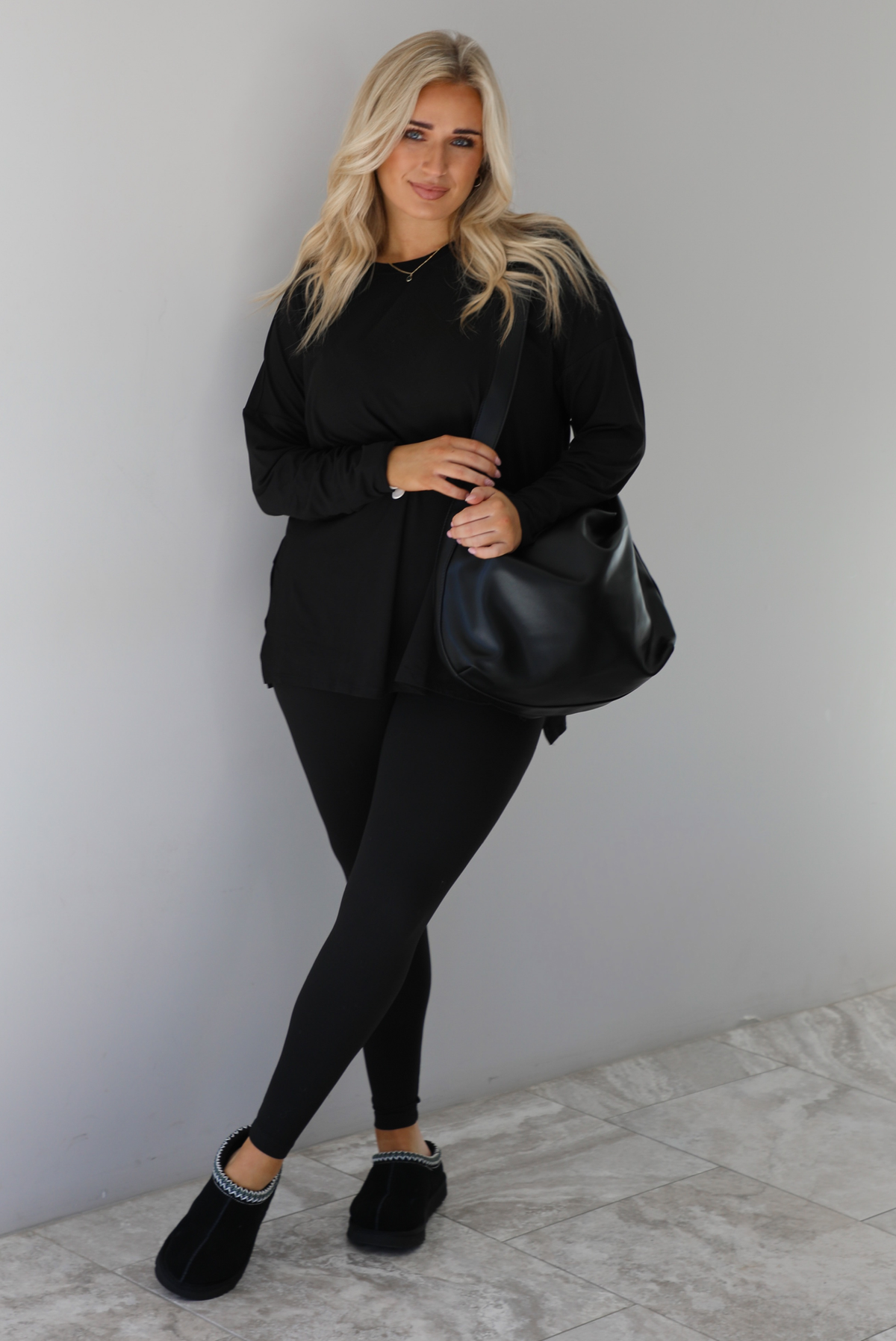 Woman wearing a black two piece set that features an oversized long sleeved top and fitted leggings. The set is paired with a black vegan leather shoulder bag and black slide on slippers. The photo is taken indoors against a white wall with white marble flooring.