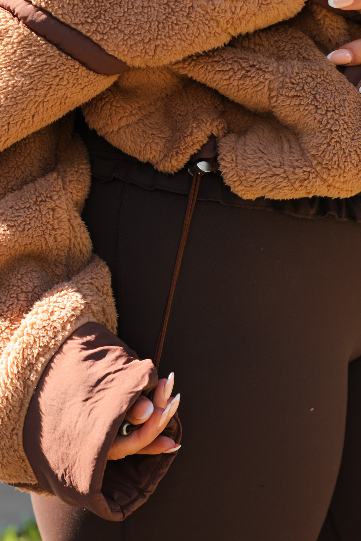 Woman model wearing a tan and brown quarter zip sweater with dark brown athleisure pants. The photo is taken outdoors against some greenery and a white wall. The photo showcases the sweater close up and the drawstring detail on the side. 