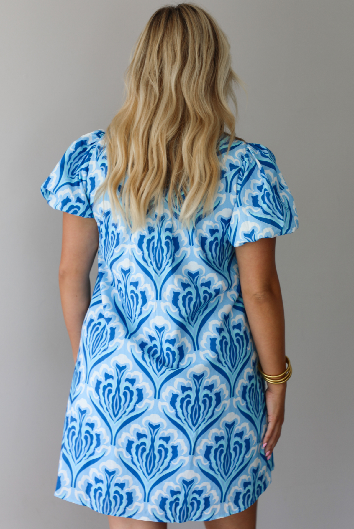 A woman modeling a short-sleeve button-up mini dress with a blue and white pattern, standing indoors against a white wall. The photo shows the back of the dress. 