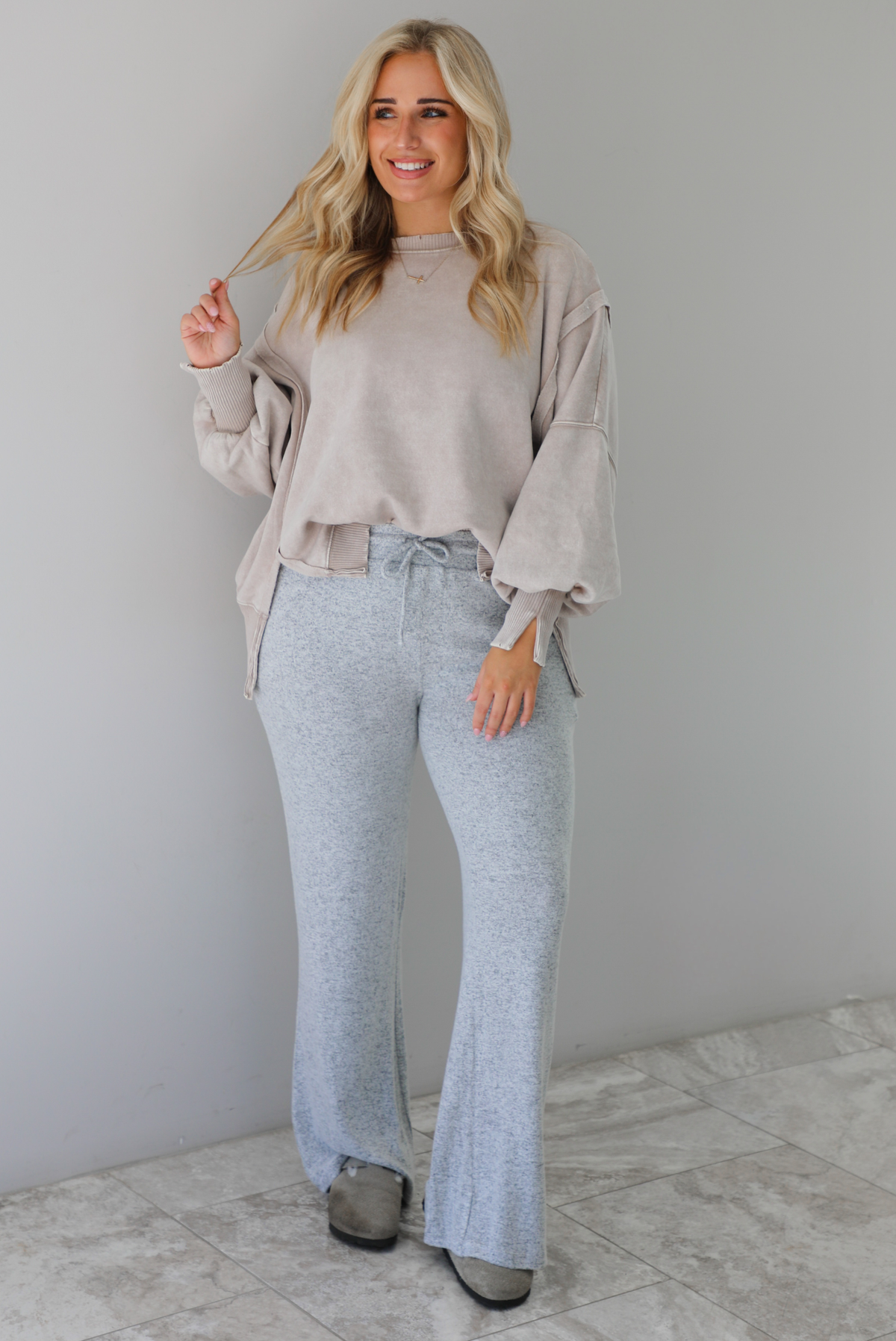 Woman wearing sand colored oversized sweatshirt with exposed seam details and a high-low design with heather grey casual pants. The outfit is styled with grey slip on shoes. The photo is taken indoors against a white wall with white marble flooring. 