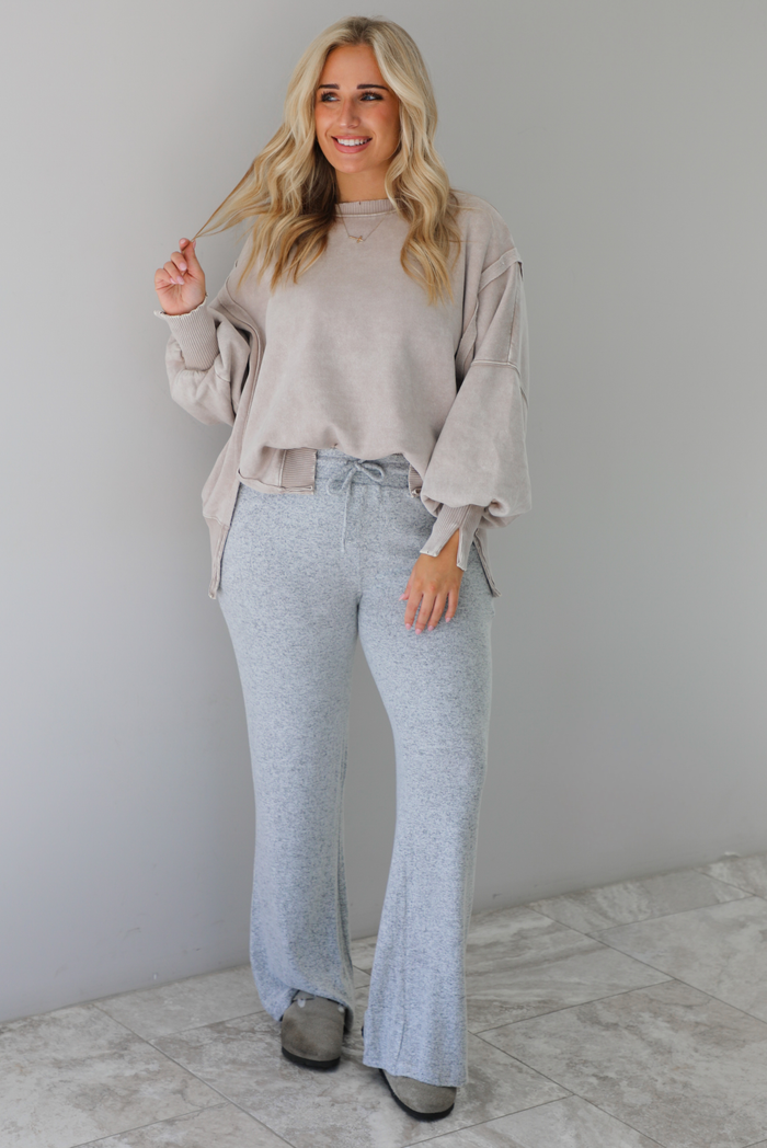 Woman wearing sand colored oversized sweatshirt with exposed seam details and a high-low design with heather grey casual pants. The outfit is styled with grey slip on shoes. The photo is taken indoors against a white wall with white marble flooring. 