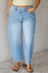 A woman wearing light-wash blue cropped barrel cut jeans with an asymmetrical button closure, standing indoors against white wall with white marble flooring. 