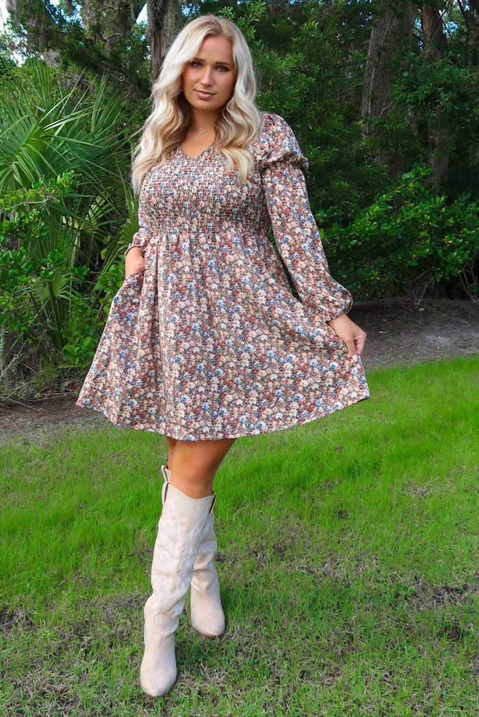 Woman model in a multi-colored floral dress and sand colored knee-high boots standing outdoors with greenery in the background. The dress has a smocked bodice, long sleeves that cinch at the wrist, and a flowy skirt that falls just above the knee on the 5'8" model. 