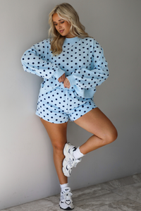 A woman wearing a blue two piece set with a navy polka dot pattern throughout, featuring a sweatshirt and high waisted sweat-shorts, paired with chunky sneakers and high ankle socks, shown indoors against a white wall with white marble flooring. 