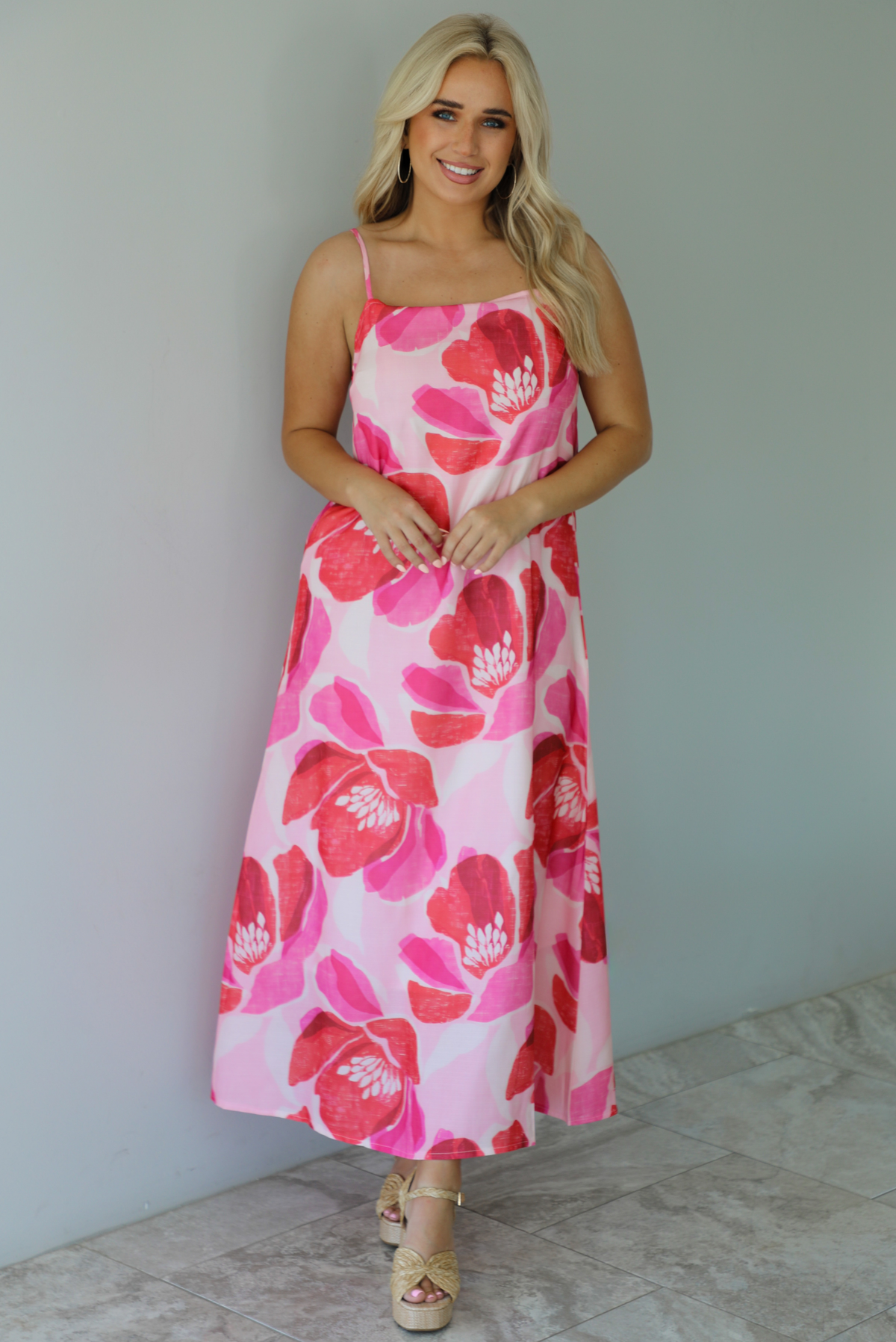 Woman wearing a pink and red floral print maxi dress with thin straps and a flowy fit indoors against a white wall with white marble flooring. 