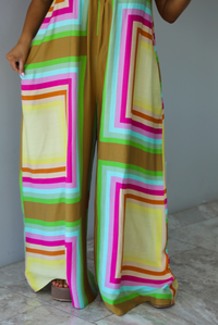 Woman wearing a colorful jumpsuit that has a high neckline, no sleeves, and a flowy cut with flared hem pants. The photo is taken indoors against a white wall with white marble flooring and shows a closeup of the flowy, flared hem pants. 