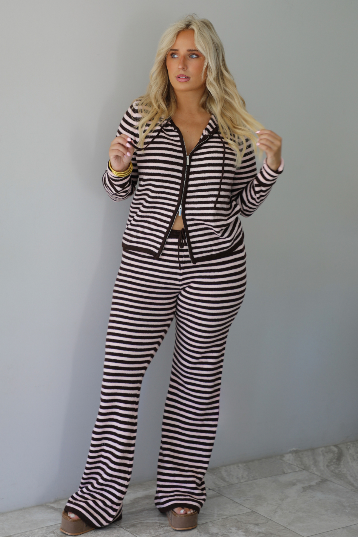 A woman wearing a light pink and brown striped two piece set featuring a double zipped hoodie top and mid-rise, wide leg pants, standing indoors against a white wall with white marble flooring. 