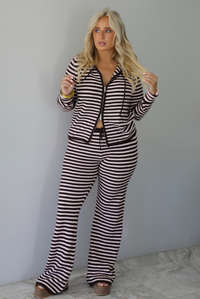 A woman wearing a light pink and brown striped two piece set featuring a double zipped hoodie top and mid-rise, wide leg pants, standing indoors against a white wall with white marble flooring. 