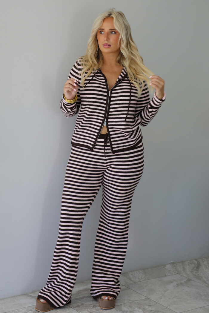A woman wearing a light pink and brown striped two piece set featuring a double zipped hoodie top and mid-rise, wide leg pants, standing indoors against a white wall with white marble flooring. 