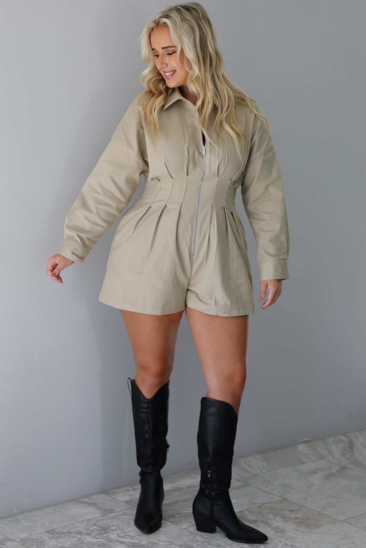 A woman wearing a sand colored long sleeved romper with a collared neckline, functional zipper closure in the front, pleated details throughout, functional pockets on either side, paired with black colored knee-high boots, standing indoors against a white wall with white marble flooring. 