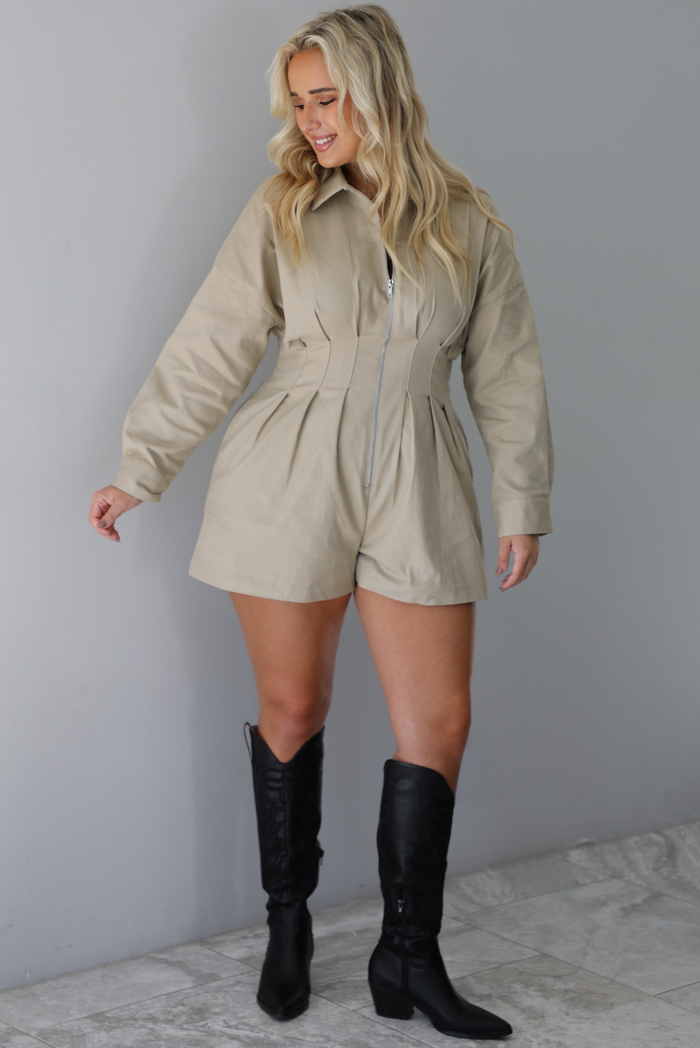 A woman wearing a sand colored long sleeved romper with a collared neckline, functional zipper closure in the front, pleated details throughout, functional pockets on either side, paired with black colored knee-high boots, standing indoors against a white wall with white marble flooring. 