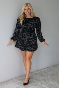 A woman wearing a black mini dress with a white polka dot print pattern throughout that has long sleeves, a high neckline, and a flowy skirt, standing indoors against a white wall with white marble flooring. 