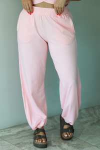 Woman wearing a pink two piece set featuring a cropped short sleeved top and high waisted jogger-style pants indoors against a white wall with white marble flooring. The photo shows a closeup of the front of the pants, which have an elastic waistline, functional pockets on either side, and a cinched detail at the ankles. 