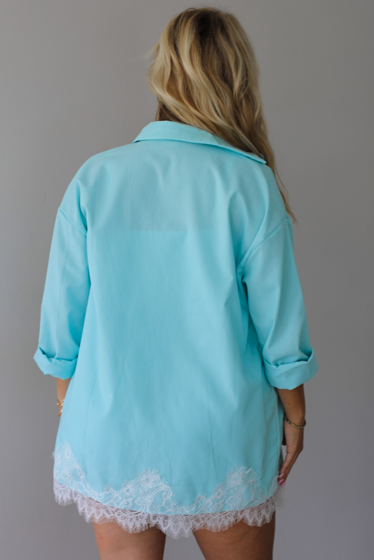 A woman stands indoors against a white wall wearing a light aqua blue matching lounge set consisting of a long open-front button-down and shorts with white lace trim, over a white tank top. The photo shows the back of the long sleeved button down. 