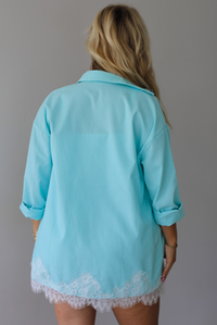 A woman stands indoors against a white wall wearing a light aqua blue matching lounge set consisting of a long open-front button-down and shorts with white lace trim, over a white tank top. The photo shows the back of the long sleeved button down. 