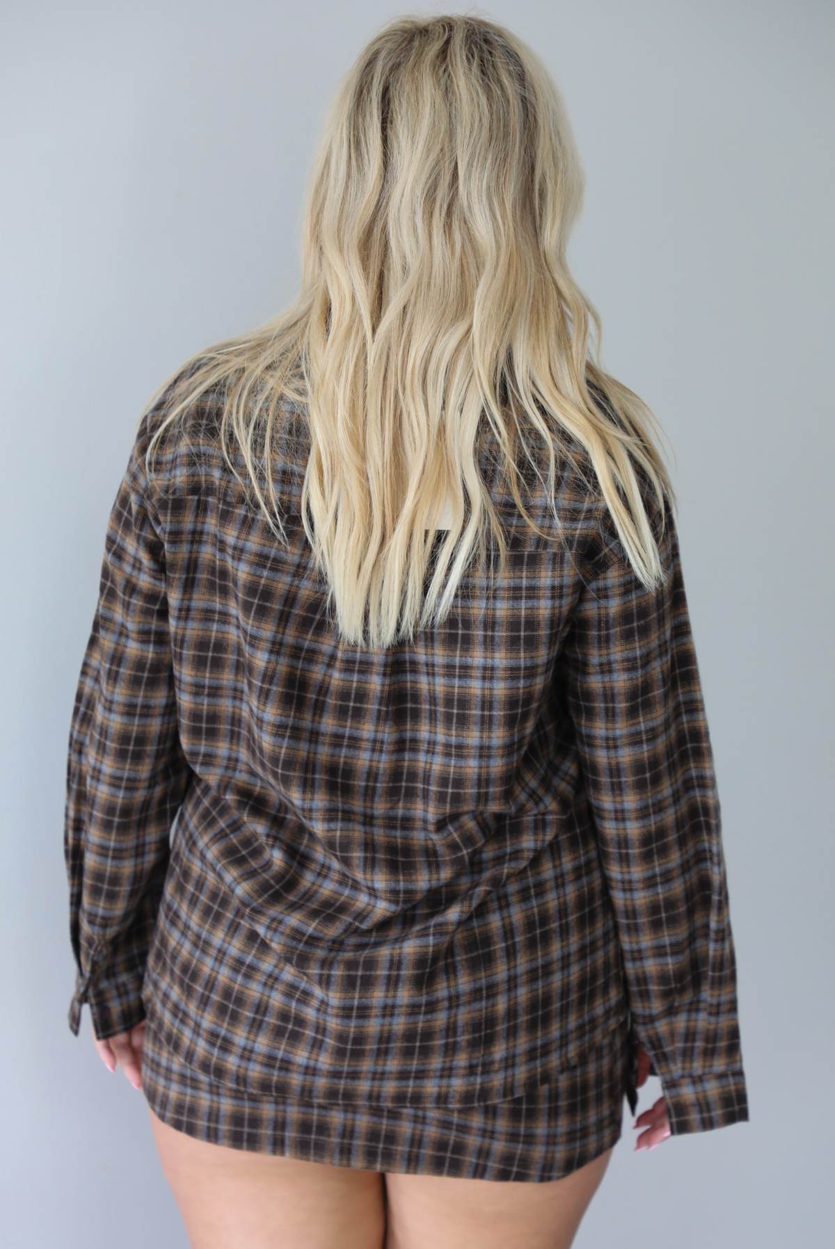 A woman shown from behind wearing aa dark brown plaid set featuring an oversized button-down and fitted mini length skirt, standing indoors against a white wall.