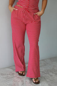 A woman modeling a red and white striped sleeveless set consisting of a tank top with non-functional button closures in the front and high waisted pants with a drawstring waistline and pockets on either side indoors against a white wall with white marble flooring. The photo shows a closeup of the pants. 
