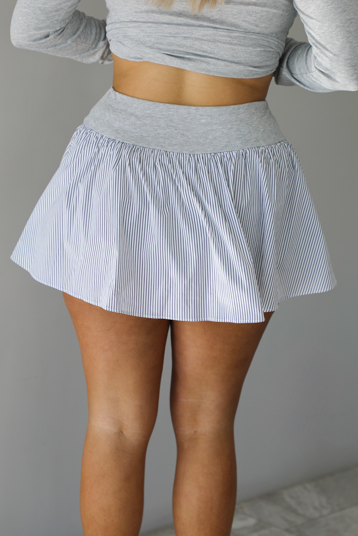 Woman wearing a grey two piece set with grey and white striped details. The top has a collared neckline, long sleeves, and functional button closures in the front. The skirt is high waisted with a flared skirt. The photo is taken indoors against a white wall and shows a closeup of the back of the skirt, which has a high waist with a ruffled, flared skirt. 