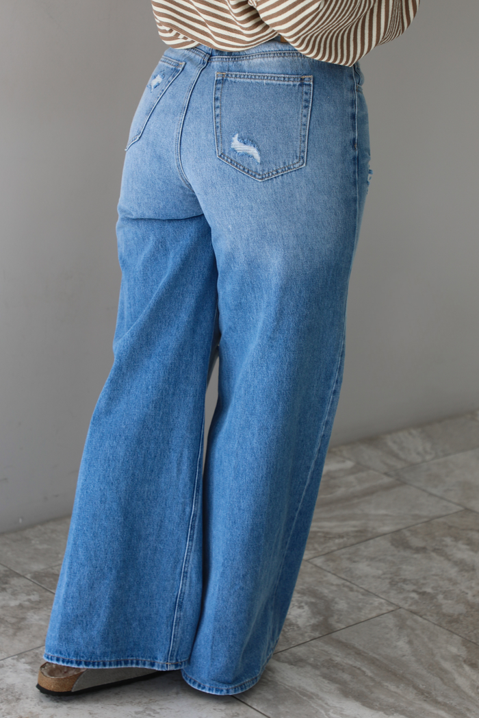 Woman wearing high waisted medium wash denim that have multiple button closures, functional belt loops, functional front and back pockets, and distressed details throughout with a flared hem. The photo shows the back of the denim. The photo is taken in front of a white wall and white marble flooring.