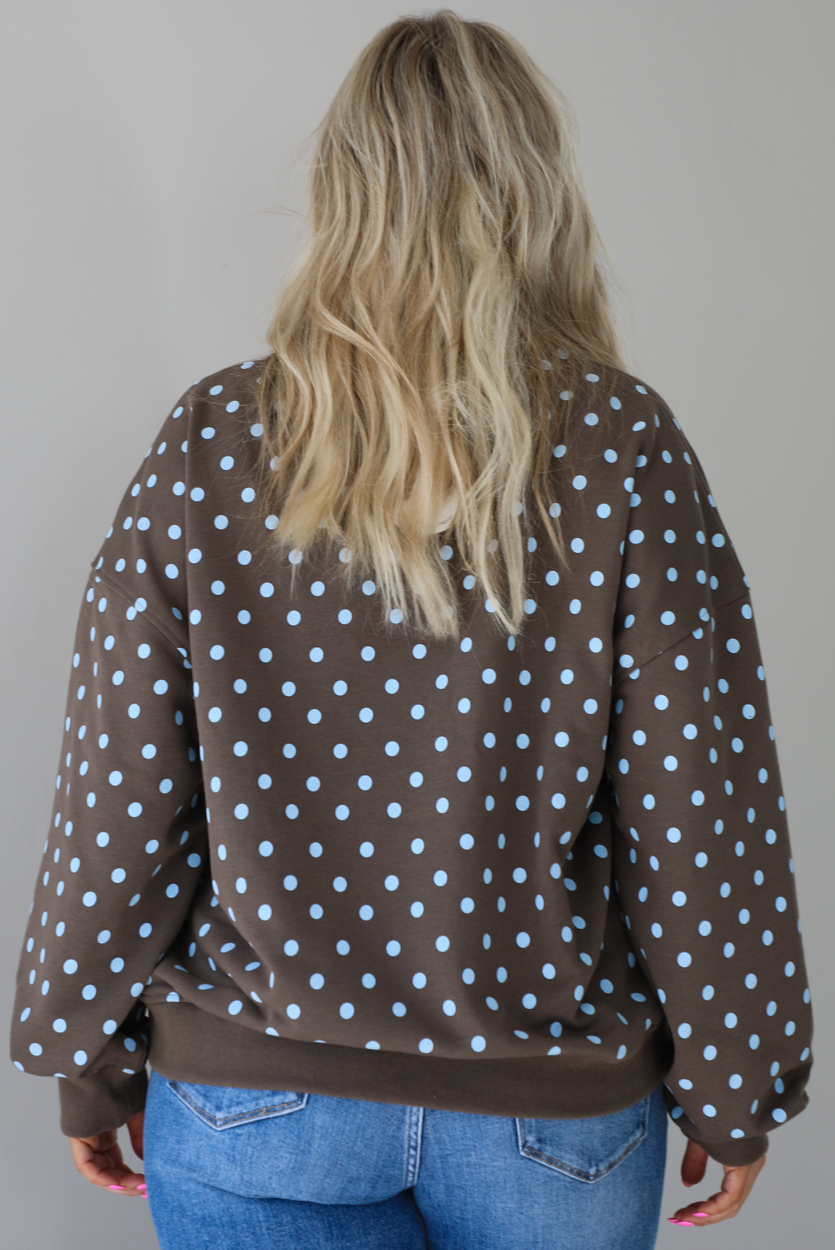Woman wearing a brown sweater that has a blue polka dot print pattern throughout with blue embroidered writing on the left side that reads 'Trust The Process.' The sweater is paired with a medium wash denim. The photo shows a closeup of the sweater, which is untucked to show the full length of the back of the sweater. The photo is taken indoors against a white wall.
