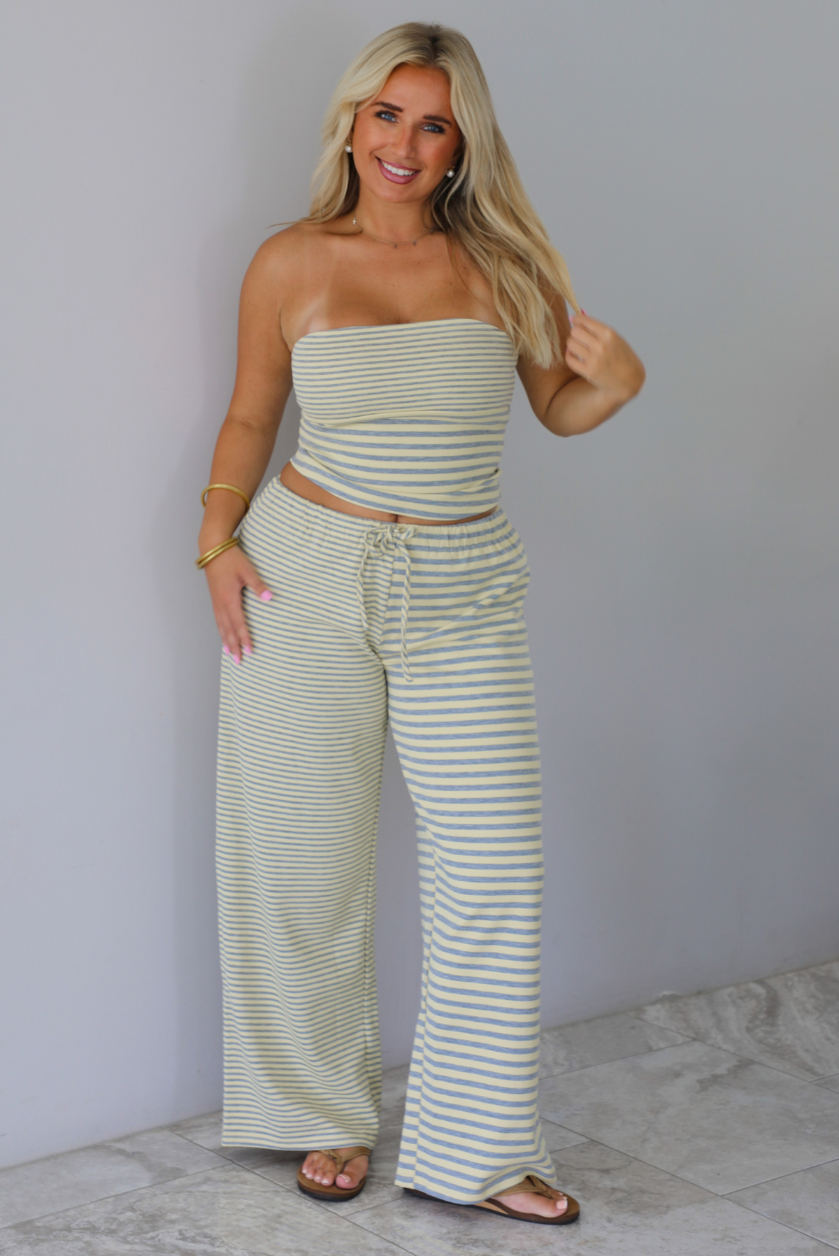 A woman wearing a pale yellow and heather grey striped two piece set with a sleeveless tube top and wide-leg pants, standing indoors against a white wall with white marble flooring. 