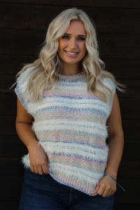 Woman model wearing a soft, striped sweater vest with dark wash denim jeans. The photo showcases a closeup of the sweater vest and the full length. The photo is taken outdoors against a dark brown wooden door. 
