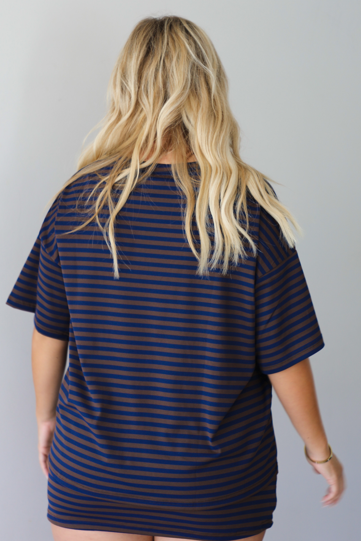 A woman shown from behind wearing a navy and brown striped set featuring an oversized top and mini skirt, standing indoors against a white wall.