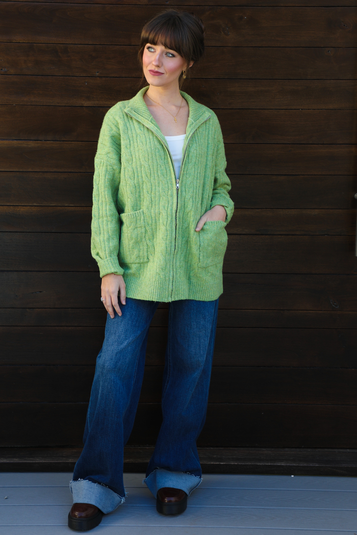 Woman model wearing a green cable knit zip-up cardigan with dark denim jeans outdoors against a dark brown wooden door. The model is wearing a Small. 