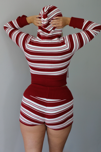 A woman shown from the back wearing a ribbed knit matching set with a hooded long-sleeve top and shorts, featuring horizontal red and white stripes against a white wall. 