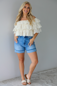 Woman wearing a cream off the shoulder, ruffled eyelet cropped top with medium wash denim shorts indoors against a white wall with white marble flooring. 