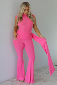 Woman wearing a bright pink jumpsuit that has a high neckline, a fitted bodice, extra fabric detail at the hips with a train on the side, and flared hem pants. The photo is taken indoors against a white wall with white marble flooring. 