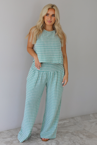 Woman wearing a mint and white gingham set with an oversized top and flared hem pants indoors against a white wall with white marble flooring. 