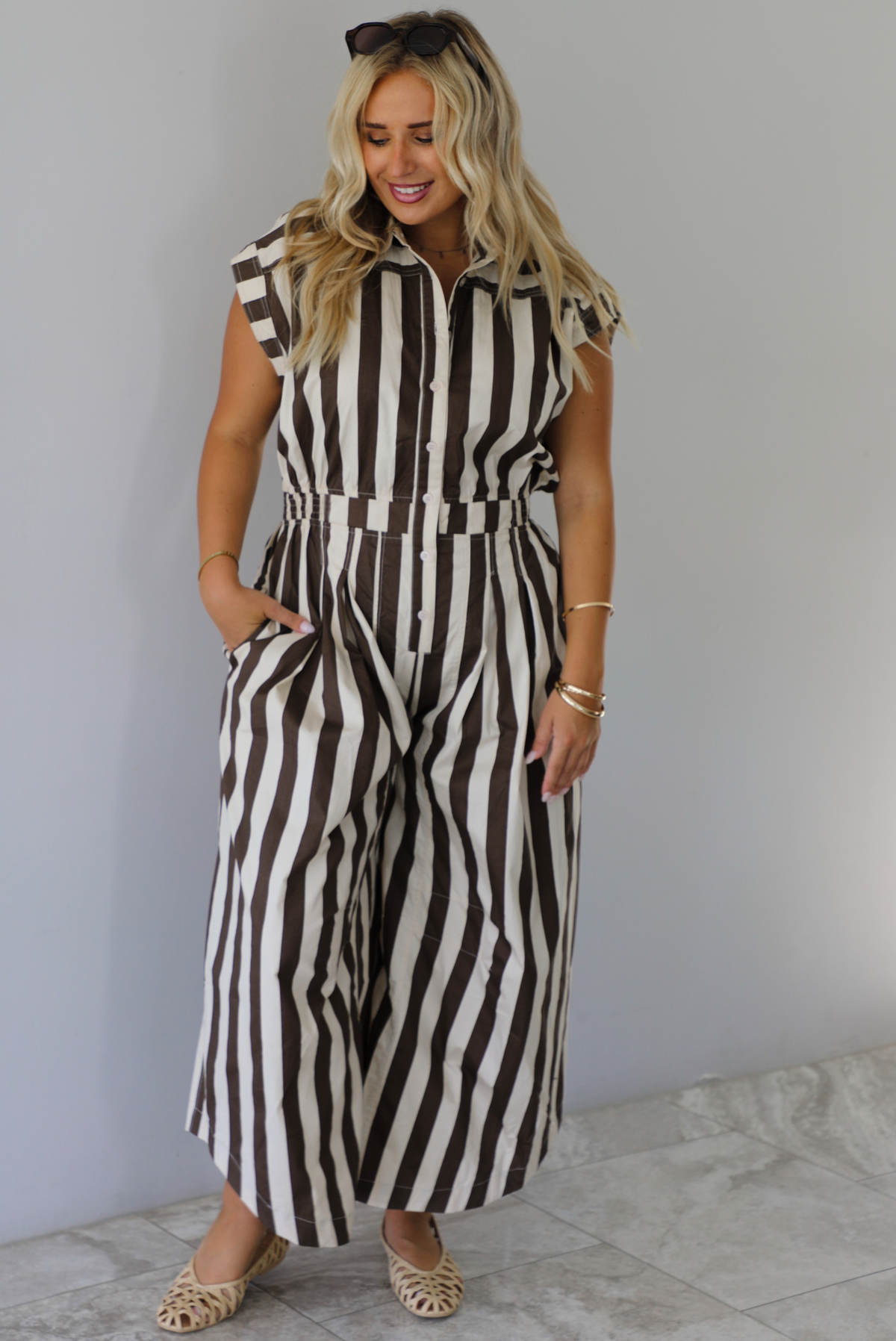 A woman wearing a brown and white striped patterned jumpsuit with a collared neckline, functional button closures in the front, a cinched waistline, and barrel-style bottoms, standing indoors against a white wall with white marble flooring. 