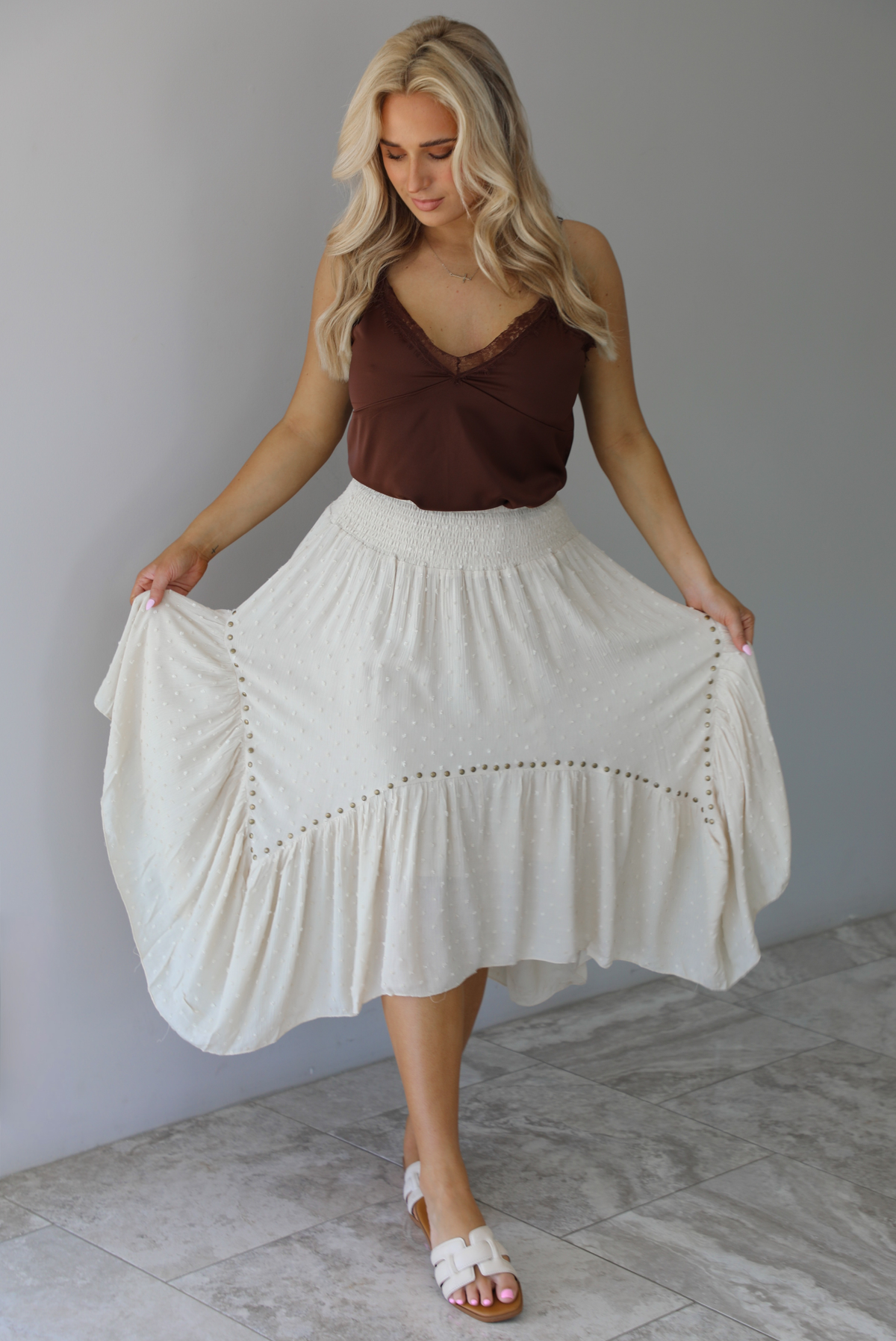 Woman wearing a satin brown tank that is tucked into a high waisted sand colored midi length skirt. The photo is taken in front of a white wall with white marble flooring. 