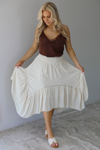 Woman wearing a satin brown tank that is tucked into a high waisted sand colored midi length skirt. The photo is taken in front of a white wall with white marble flooring. 