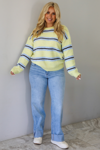 Woman wearing a pastel yellow, blue, and dark brown striped sweater, paired with light wash denim jeans that have a wide leg cut with a cuffed hem, standing indoors against a white wall with white marble flooring.