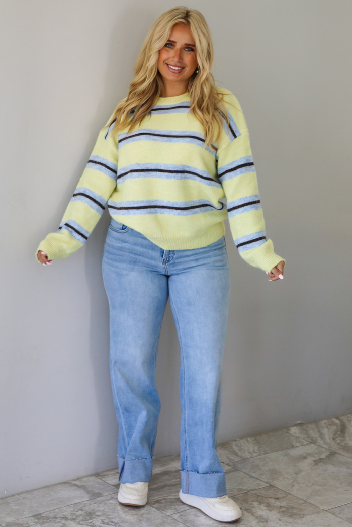 Woman wearing a pastel yellow, blue, and dark brown striped sweater, paired with light wash denim jeans that have a wide leg cut with a cuffed hem, standing indoors against a white wall with white marble flooring.