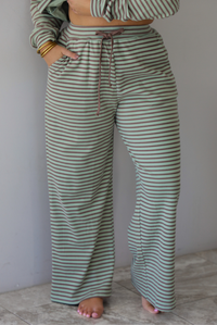 A woman wearing a pale green and brown striped two piece set featuring an oversized long sleeved top and high waisted, wide leg pants, standing indoors against a white wall with white marble flooring. The photo shows a closeup of the pants, which have an elastic and drawstring waistline and functional pockets on either side.